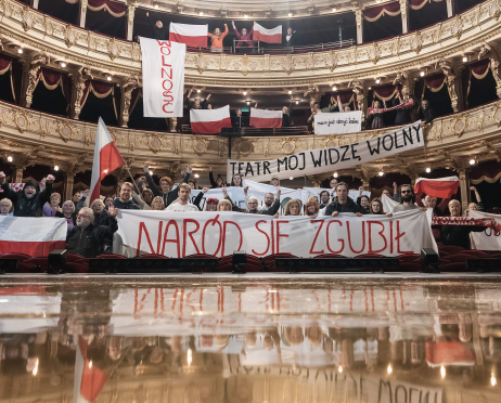 Grupa osób protestujących zgromadzonych w zdobionym wnętrzu teatru. Uczestnicy stoją na parterze i balkonach, trzymając biało-czerwone flagi oraz transparenty z hasłami, wielu z nich unosi pięści w geście sprzeciwu. Na pierwszym planie widoczny jest duży baner z napisem „Naród się zgubił”. W tle złocone balkony i czerwone kurtyny.